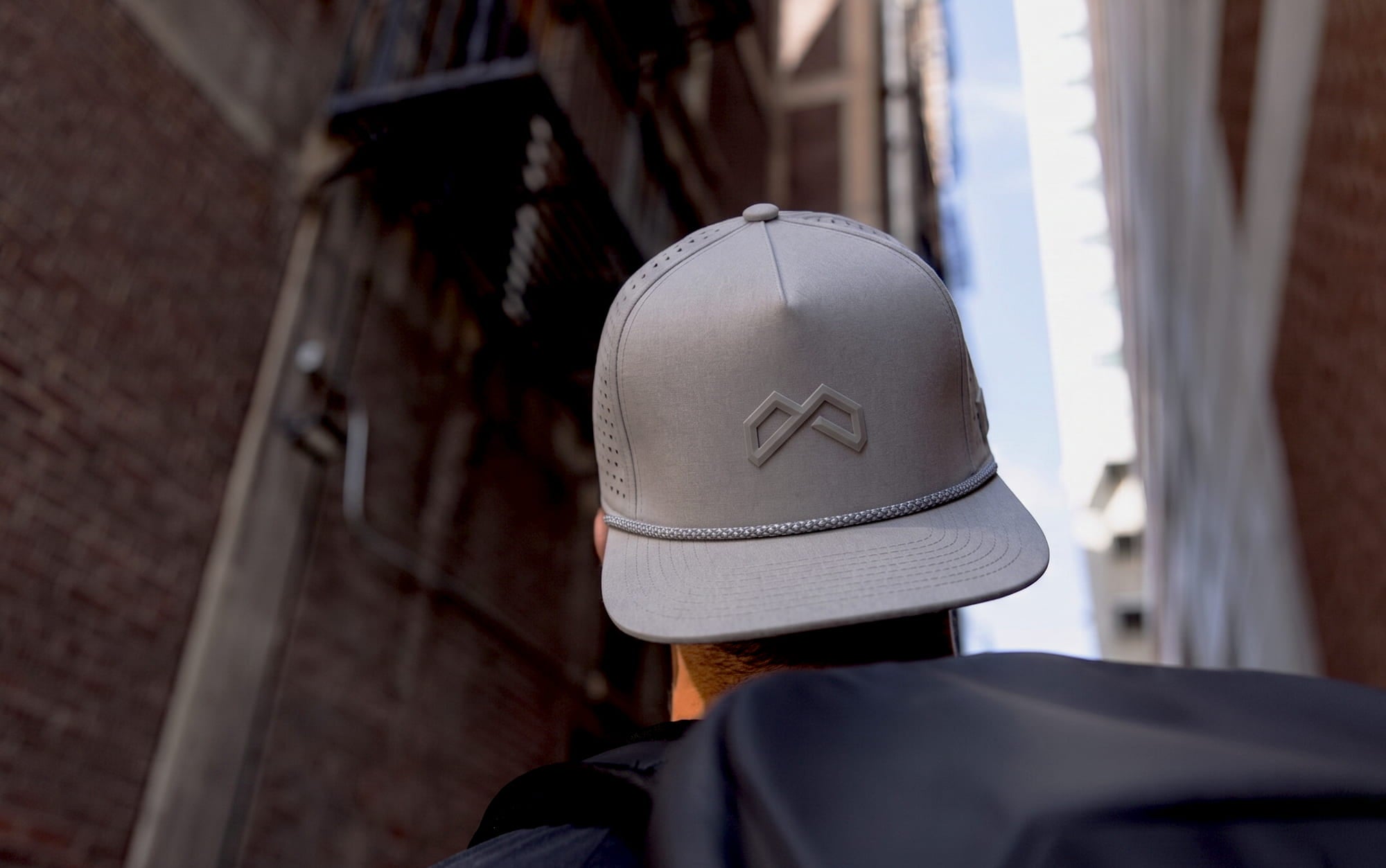 Person seen from behind wearing a gray flat-brim cap with a raised mountain logo in a narrow urban alley.